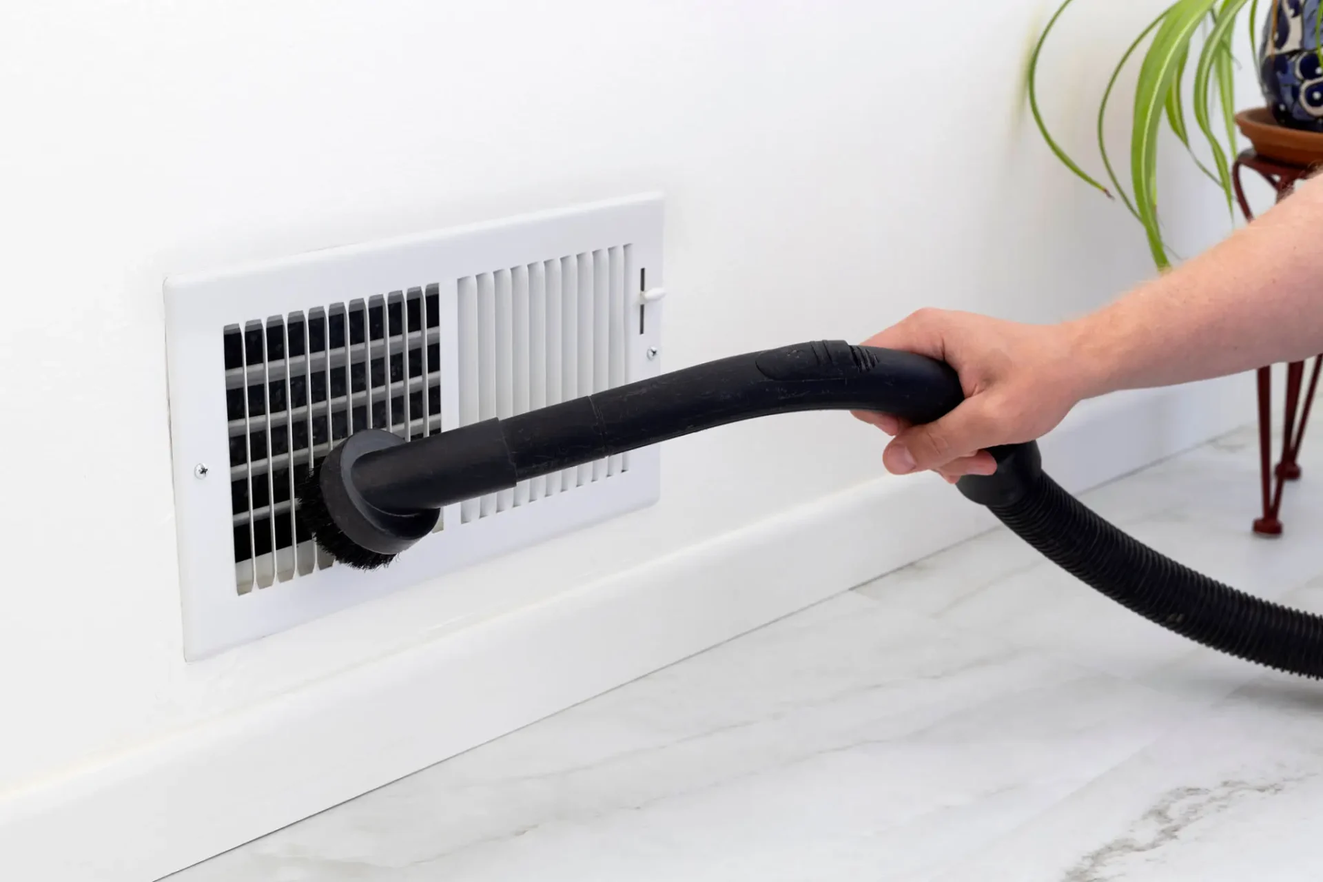 Person vacuuming dust from an air vent in a white room.